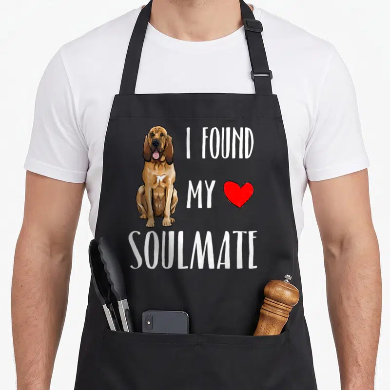 Found dog apron - Cartoon brown bloodhound sits left of white text I Found My Soulmate and red heart on grey background — shown on a black cotton canvas apron against a white background