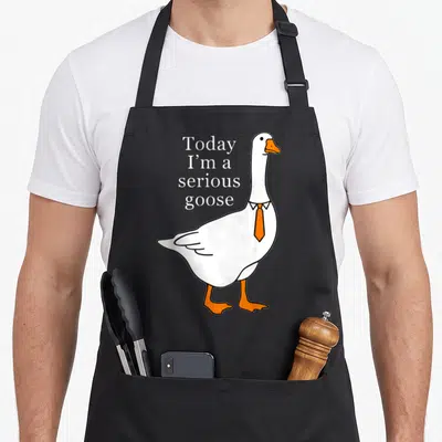 Serious goose apron - Cartoon white goose with orange beak and tie next to stacked white text — shown on a black cotton canvas apron against a white background