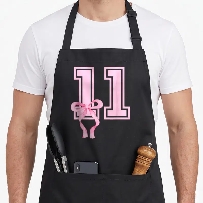 Birthday coquette bow apron - Pink number 11 with watercolor bow overlapping the first digit — shown on a black cotton canvas apron against a white background