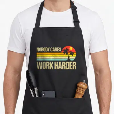Motivational work hard apron - Retro sunset graphic with horizontal stripes and two palm tree silhouettes between — shown on a black cotton canvas apron against a white background