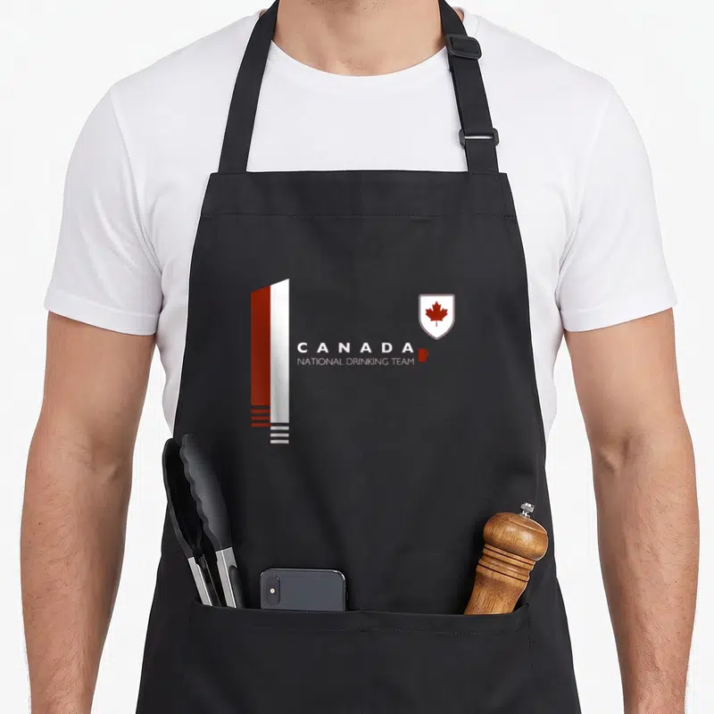 Canada beer apron - Red white vertical bar, CANADA NATIONAL DRINKING TEAM text, maple leaf shield, small mug icon — shown on a black cotton canvas apron against a white background
