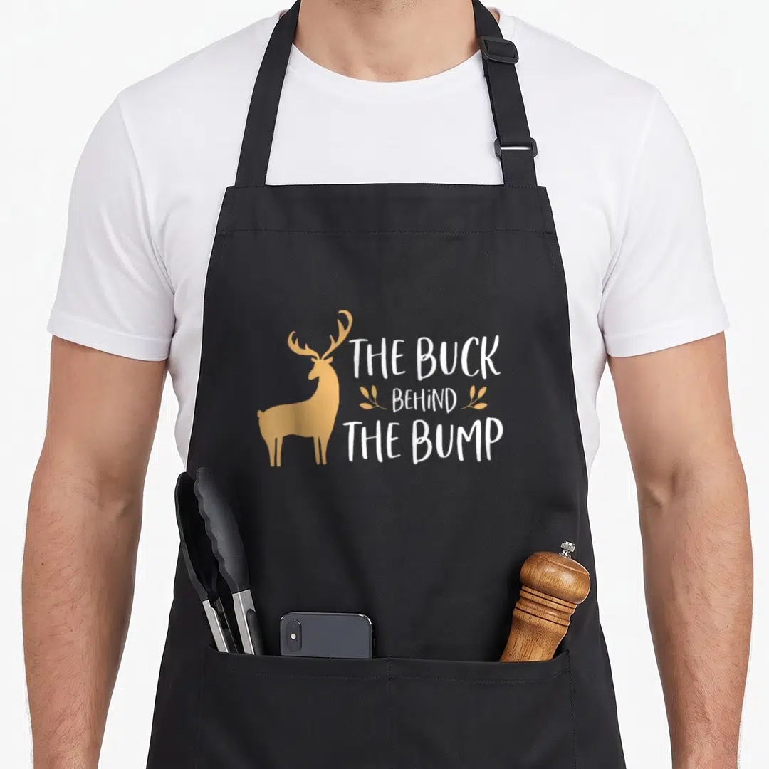 Pregnancy christmas apron - Tan deer silhouette next to white text THE BUCK BEHIND THE BUMP with small leaf sprigs — shown on a black cotton canvas apron against a white background