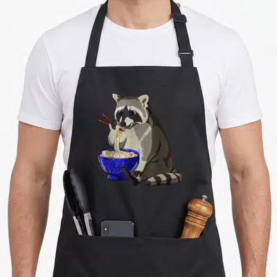 Raccoon anime apron - Cartoon raccoon holding red chopsticks eating yellow noodles from a blue patterned bowl — shown on a black cotton canvas apron against a white background