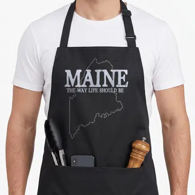 Maine pride apron - White Maine state outline with slogan MAINE THE WAY LIFE SHOULD BE — shown on a black cotton canvas apron against a white background