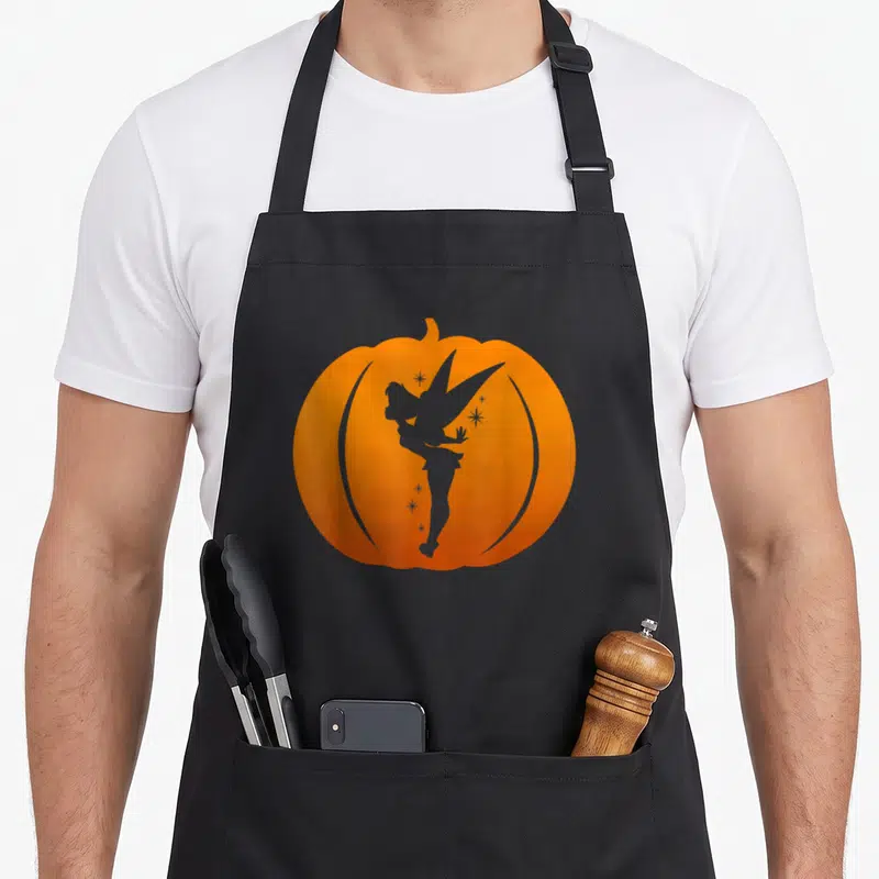 Tinker halloween kitchen aprons - Orange pumpkin silhouette containing a fairy shape and scattered stars — shown on a black cotton canvas apron against a white background