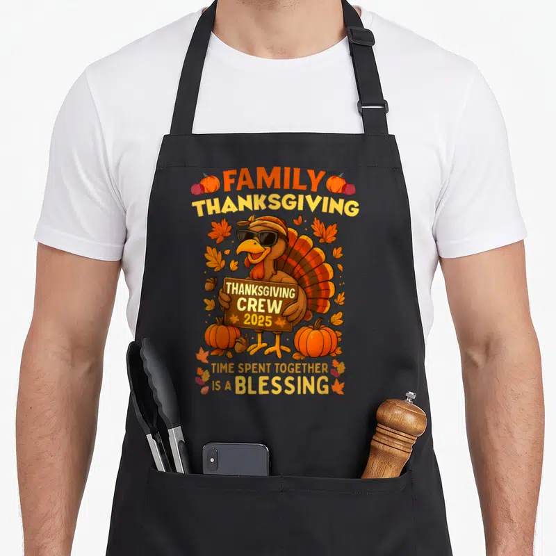 Crew thanksgiving aprons - Cartoon turkey with sunglasses holding 2025 sign surrounded by pumpkins and autumn leaves — shown on a black cotton canvas apron against a white background