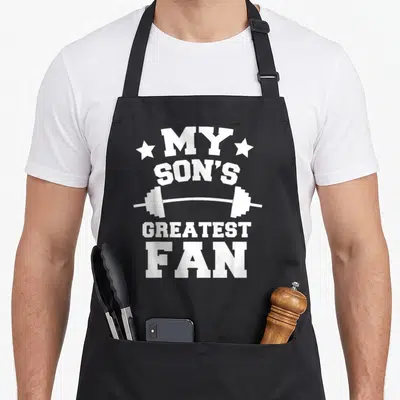 Powerlifting dad and son aprons - White text My Son's Greatest Fan separated by a barbell illustration with stars — shown on a black cotton canvas apron against a white background