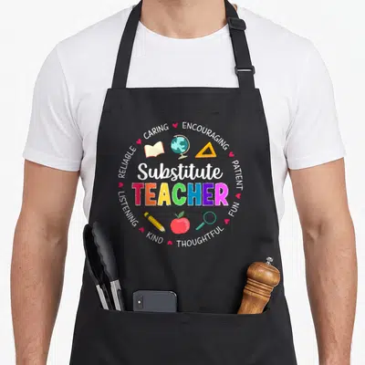 Substitute appreciation teacher apron - Circular design with Substitute TEACHER text, school icons, and adjectives like — shown on a black cotton canvas apron against a white background