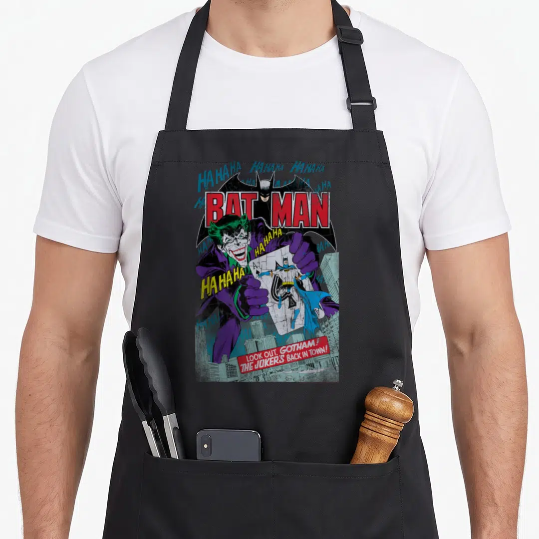Funny cover apron - Joker holding Batman playing card with Gotham skyline and scattered laughter text — shown on a black cotton canvas apron against a white background