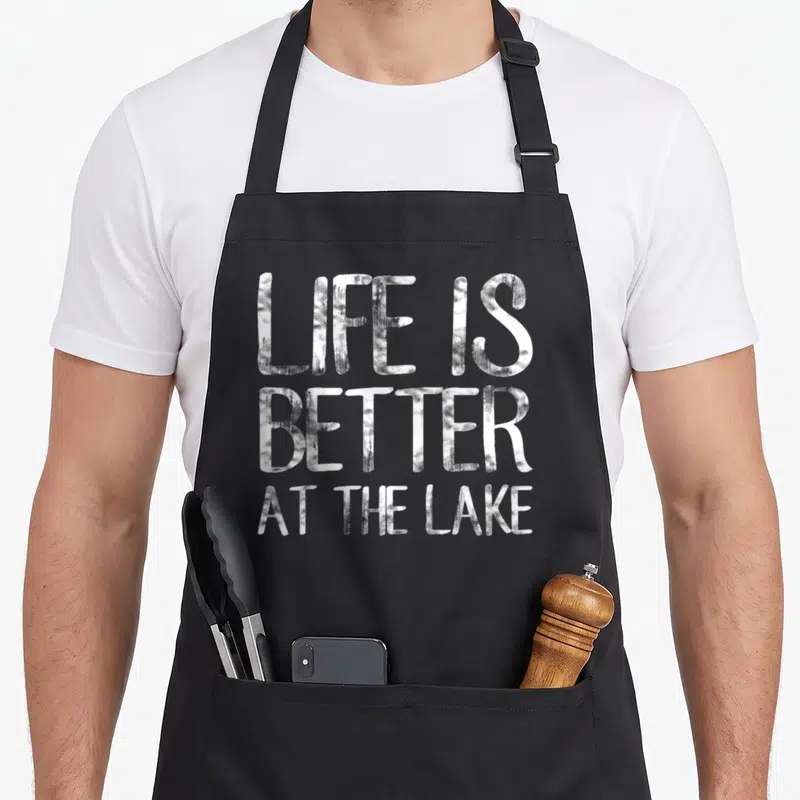 Better fishing aprons - White distressed typography reading LIFE IS BETTER AT THE LAKE stacked vertically — shown on a black cotton canvas apron against a white background