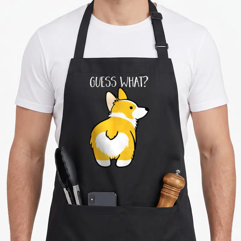 Butt dog apron - Cartoon corgi facing away with GUESS WHAT? text in orange and white — shown on a black cotton canvas apron against a white background