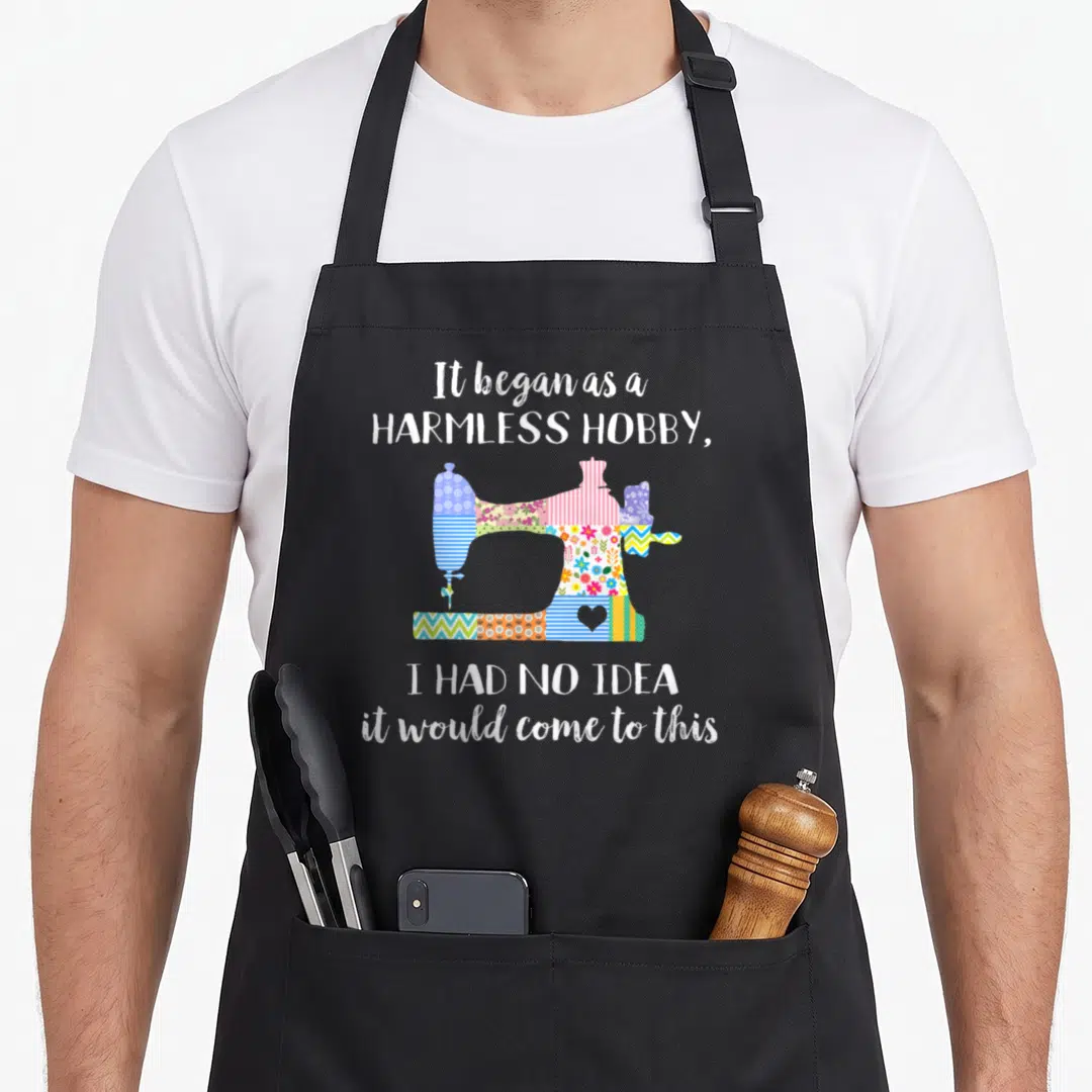Quilter hobby apron - Cartoon sewing machine filled with colorful fabric patterns between white text phrases — shown on a black cotton canvas apron against a white background