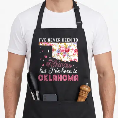 Heaven oklahoma boy apron - Floral Oklahoma state map with stacked text and scattered pink hearts and stars — shown on a black cotton canvas apron against a white background