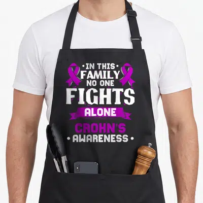 Inflammatory crohns awareness apron - White and purple text reading IN THIS FAMILY NO ONE FIGHTS ALONE with two — shown on a black cotton canvas apron against a white background