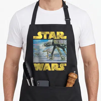 Imperial at apron - Yellow Star Wars text above and below rectangular AT-AT walker illustration on snow — shown on a black cotton canvas apron against a white background