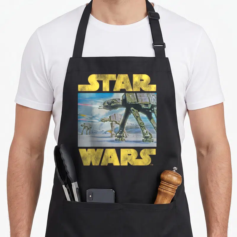 Imperial at apron - Yellow Star Wars text above and below rectangular AT-AT walker illustration on snow — shown on a black cotton canvas apron against a white background