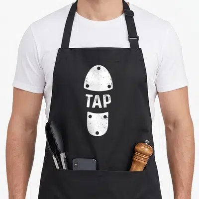 Stepptanzer shoe apron - White TAP text between two vertical shoe soles with rivet holes — shown on a black cotton canvas apron against a white background