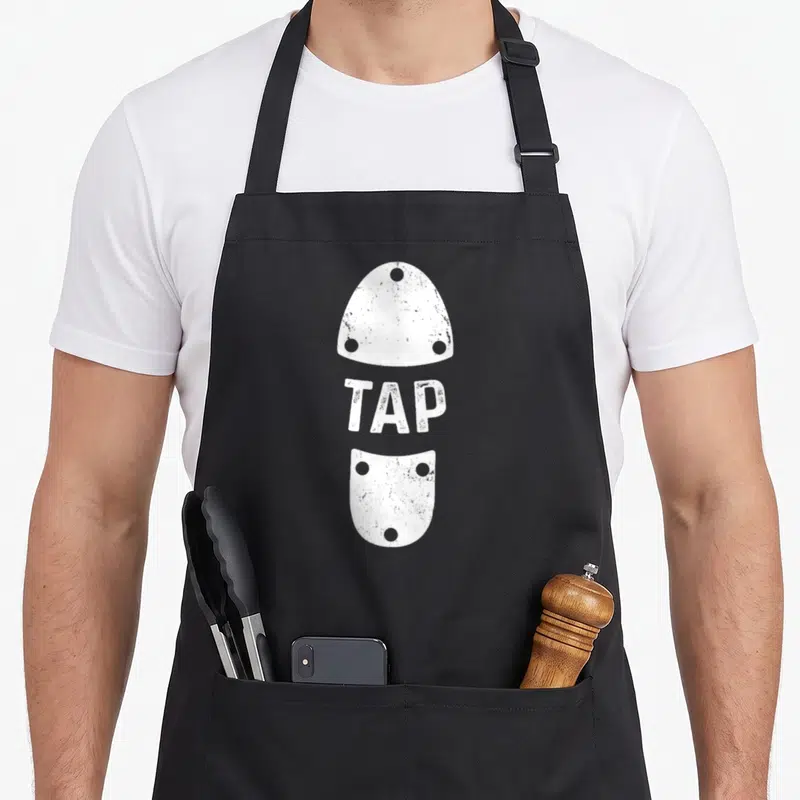 Stepptanzer shoe apron - White TAP text between two vertical shoe soles with rivet holes — shown on a black cotton canvas apron against a white background
