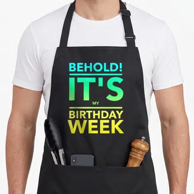 Funny week birthday apron - Stacked cyan green and yellow text reading BEHOLD IT'S MY BIRTHDAY WEEK separated — shown on a black cotton canvas apron against a white background