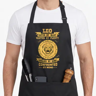 Astrology zodiac aprons - Yellow distressed lion head in circle with banners reading HATED BY MANY WANTED BY PLENTY — shown on a black cotton canvas apron against a white background