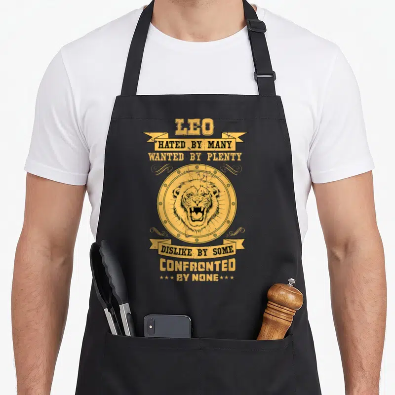 Astrology zodiac aprons - Yellow distressed lion head in circle with banners reading HATED BY MANY WANTED BY PLENTY — shown on a black cotton canvas apron against a white background