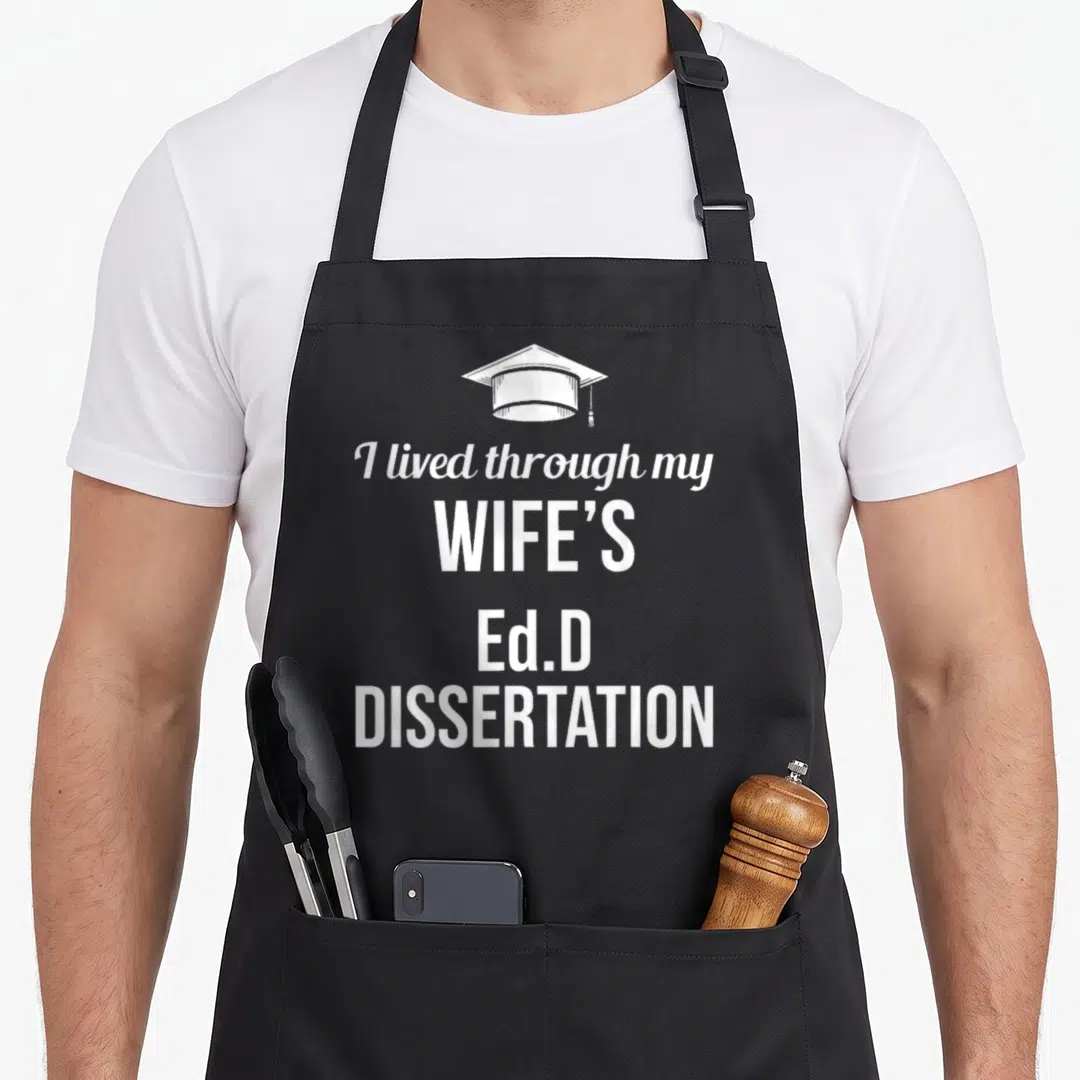 Wife doctor apron - White graduation cap above stacked text reading I lived through my wife s Ed.D dissertation — shown on a black cotton canvas apron against a white background