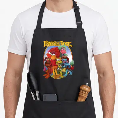 Fraggle rock apron - Stacked yellow title above red, blue, green, and yellow furry characters huddled in a blue — shown on a black cotton canvas apron against a white background