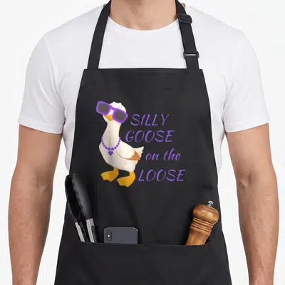 Funny goose silly apron - Cartoon white goose in purple sunglasses and necklace beside stacked purple text — shown on a black cotton canvas apron against a white background