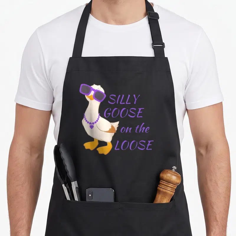 Funny goose silly apron - Cartoon white goose in purple sunglasses and necklace beside stacked purple text — shown on a black cotton canvas apron against a white background