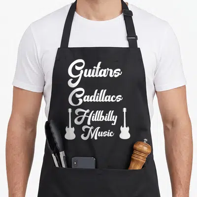 Hillbilly music cook's country aprons - White stacked text Guitars Cadillacs Hillbilly Music flanked by two guitar — shown on a black cotton canvas apron against a white background