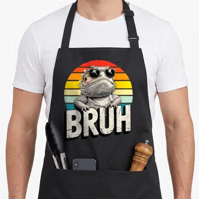 Gaming dragon apron - Bearded dragon with sunglasses and BRUH text over retro striped sun — shown on a black cotton canvas apron against a white background