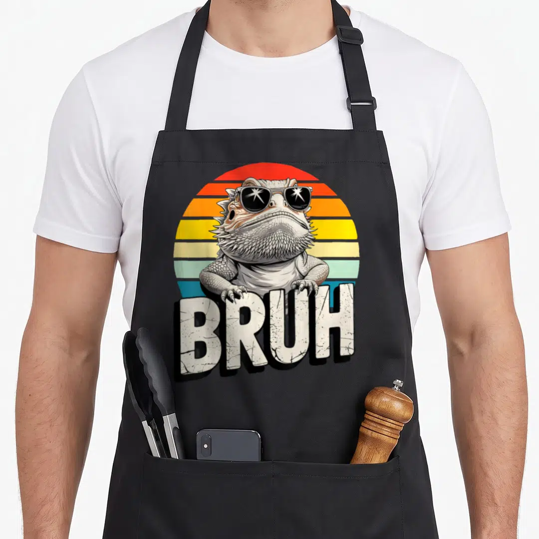 Gaming dragon apron - Bearded dragon with sunglasses and BRUH text over retro striped sun — shown on a black cotton canvas apron against a white background