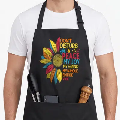 Sunflower colorful aprons - Stylized sunflower with stacked text reading DON'T DISTURB my PEACE MY JOY and small symbols — shown on a black cotton canvas apron against a white background