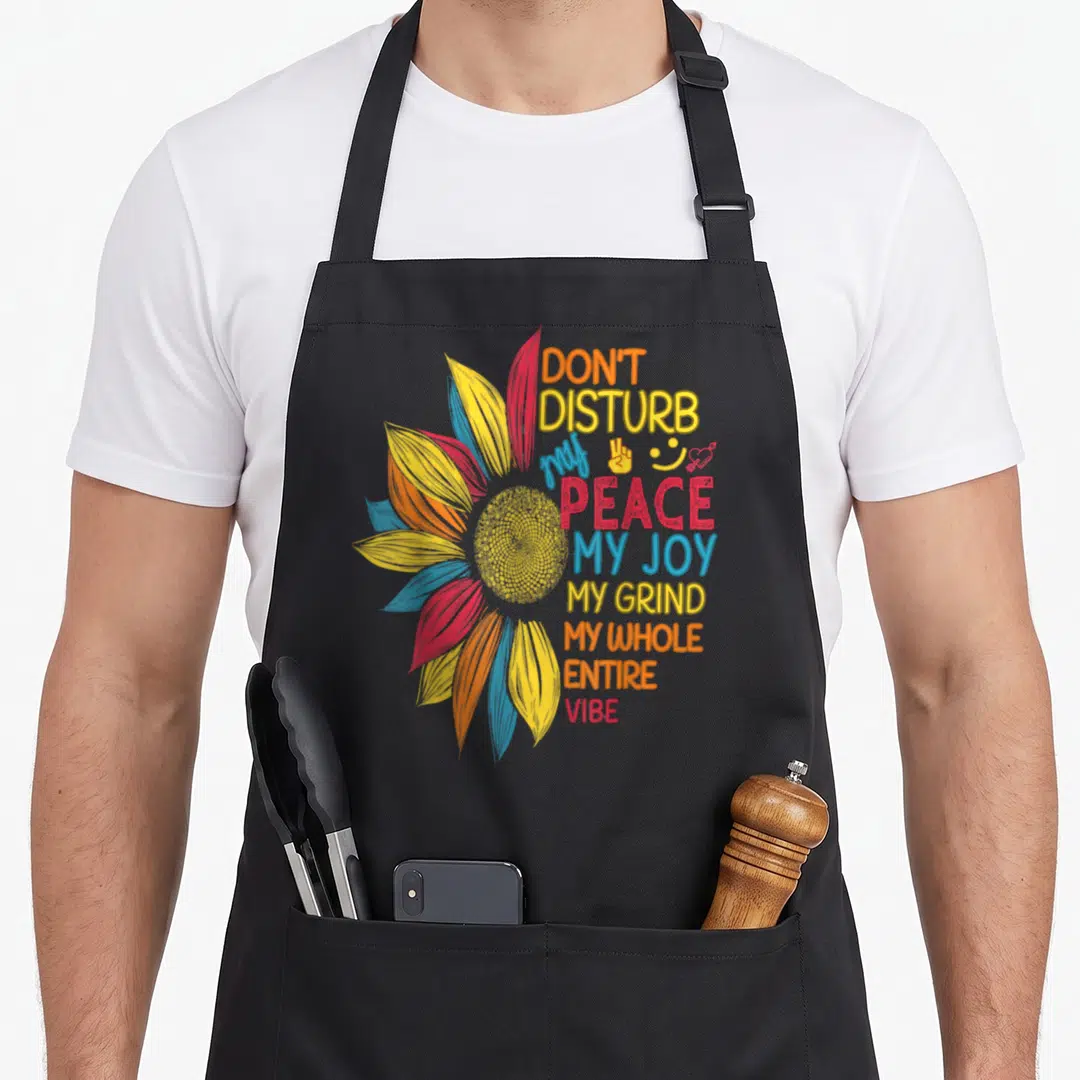 Sunflower colorful aprons - Stylized sunflower with stacked text reading DON'T DISTURB my PEACE MY JOY and small symbols — shown on a black cotton canvas apron against a white background