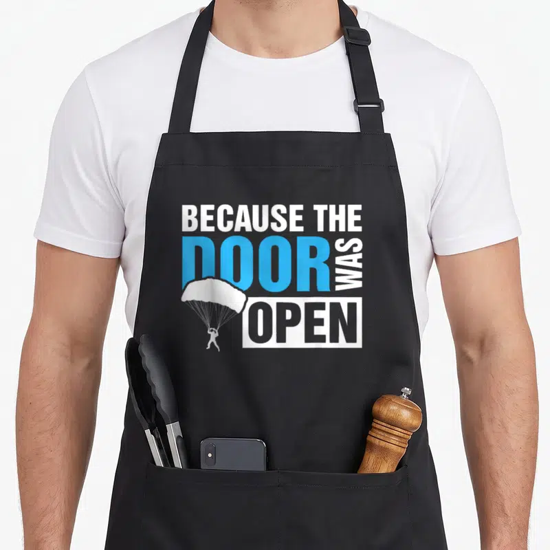 Open door apron - Skydiver silhouette overlapping stacked white and blue text reading Because The Door Was Open — shown on a black cotton canvas apron against a white background