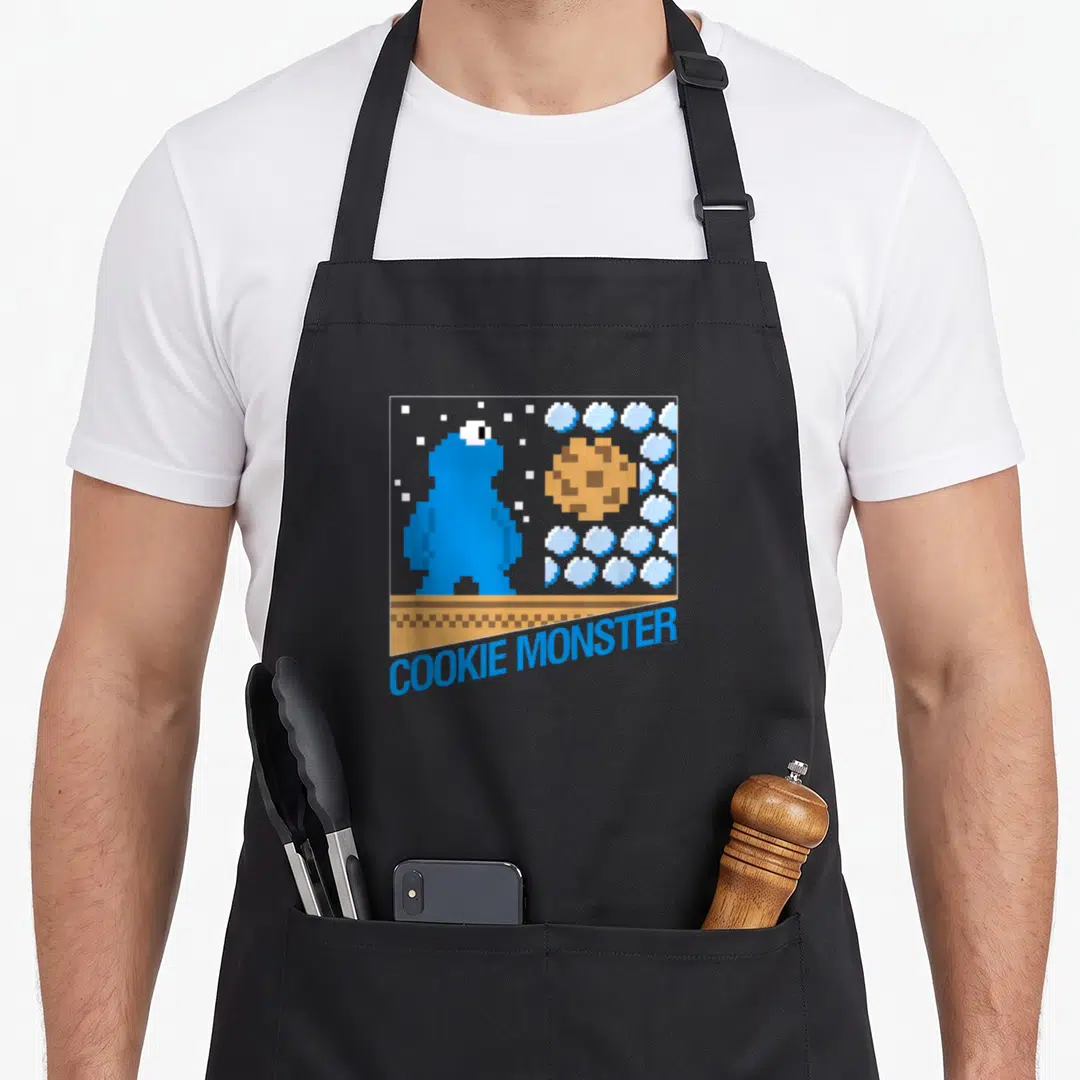 Bit cookie monster apron - Blue pixel character facing brown cookie in square frame with white dots and blue text below — shown on a black cotton canvas apron against a white background