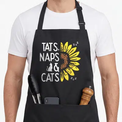 Tat cat apron - Distressed text TATS NAPS CATS with half sunflower and Zzz symbols — shown on a black cotton canvas apron against a white background