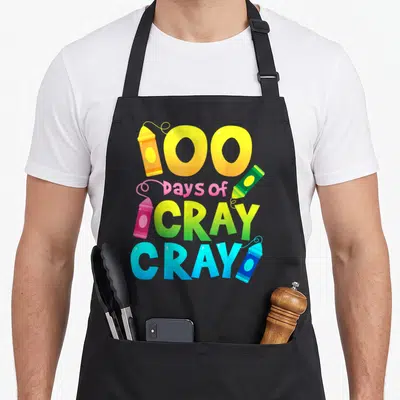 Cray school teacher apron - Stacked 100 days of CRAY CRAY text with four yellow pink green and blue cartoon crayons — shown on a black cotton canvas apron against a white background