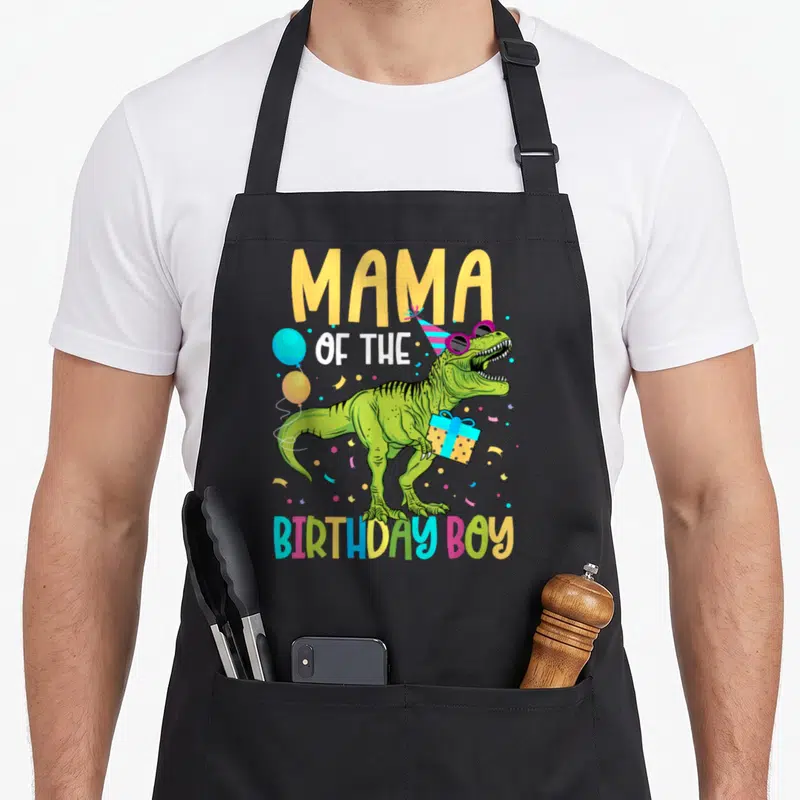 Troop mama apron - Green T-Rex wearing sunglasses and party hat holding a gift under colorful text — shown on a black cotton canvas apron against a white background
