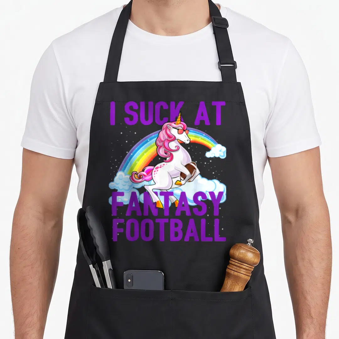 Suck rainbow apron - Cartoon unicorn holding football under rainbow with I Suck At Fantasy Football text — shown on a black cotton canvas apron against a white background