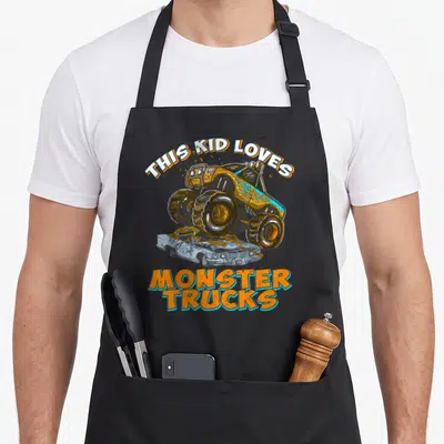 Monster pickup truck apron - THIS KID LOVES MONSTER TRUCKS text with orange and blue truck crushing silver car — shown on a black cotton canvas apron against a white background