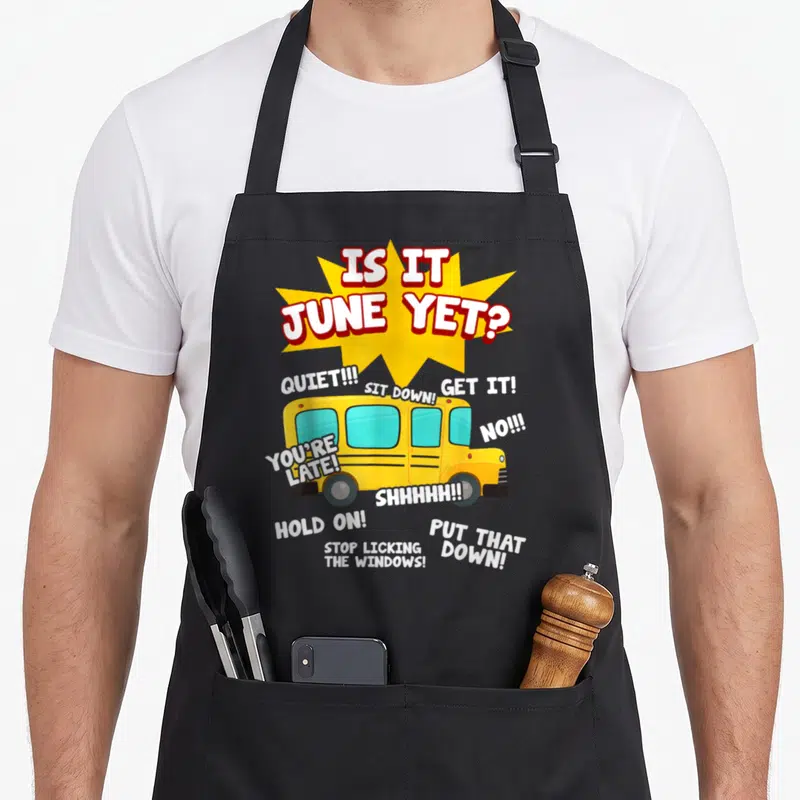 June student aprons - Yellow school bus with IS IT JUNE YET text and scattered command phrases like SIT DOWN and STOP — shown on a black cotton canvas apron against a white background