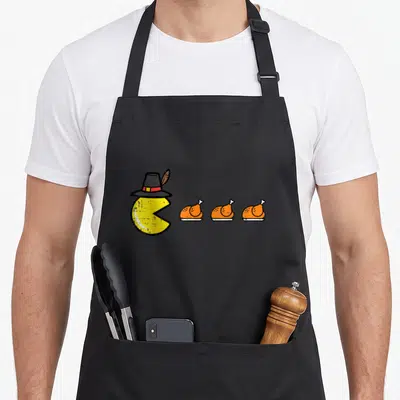 Video turkey apron - Yellow Pac-Man in black pilgrim hat facing three orange roasted turkeys on white plates — shown on a black cotton canvas apron against a white background
