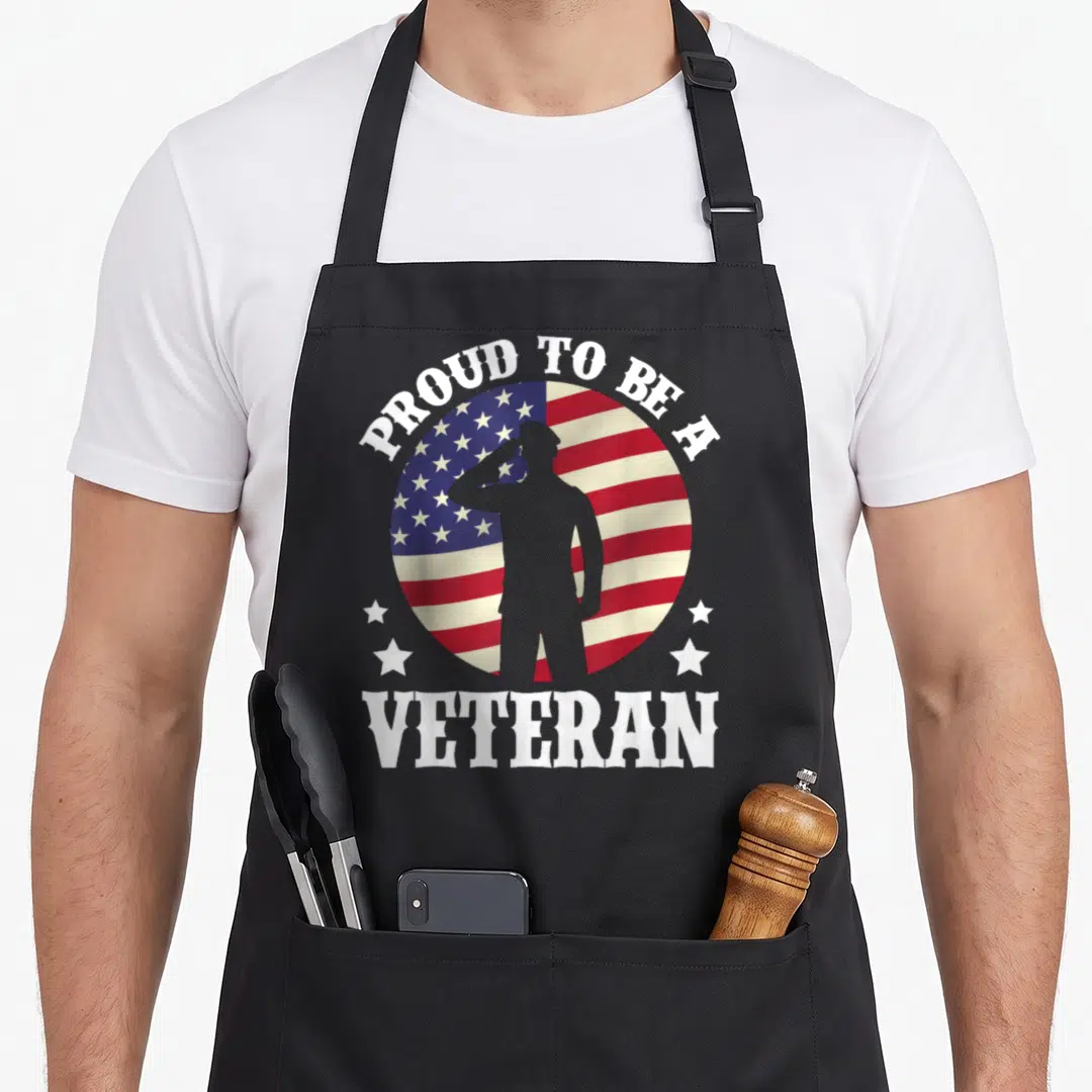 Proud veterans day apron - Saluting soldier silhouette on circular flag with Proud To Be A Veteran text — shown on a black cotton canvas apron against a white background