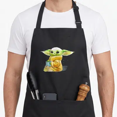 Illustration soup apron - Watercolor Grogu seated holding a blue bowl with green contents — shown on a black cotton canvas apron against a white background