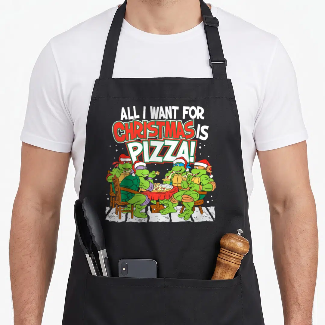 Christmas pizza apron - Four cartoon turtles in Santa hats eating pizza under stacked text All I Want For Christmas Is — shown on a black cotton canvas apron against a white background