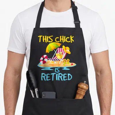 Chick retirement apron - Cartoon chick in beach chair with THIS CHICK IS RETIRED text — shown on a black cotton canvas apron against a white background