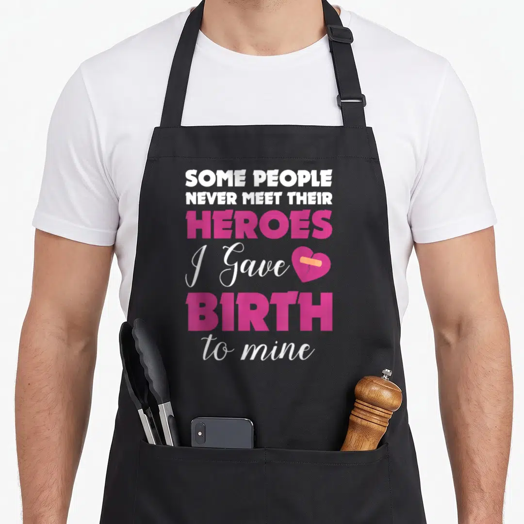 Awareness congenital heart defect apron - Stacked white and pink text reading heroes I gave birth to mine next — shown on a black cotton canvas apron against a white background