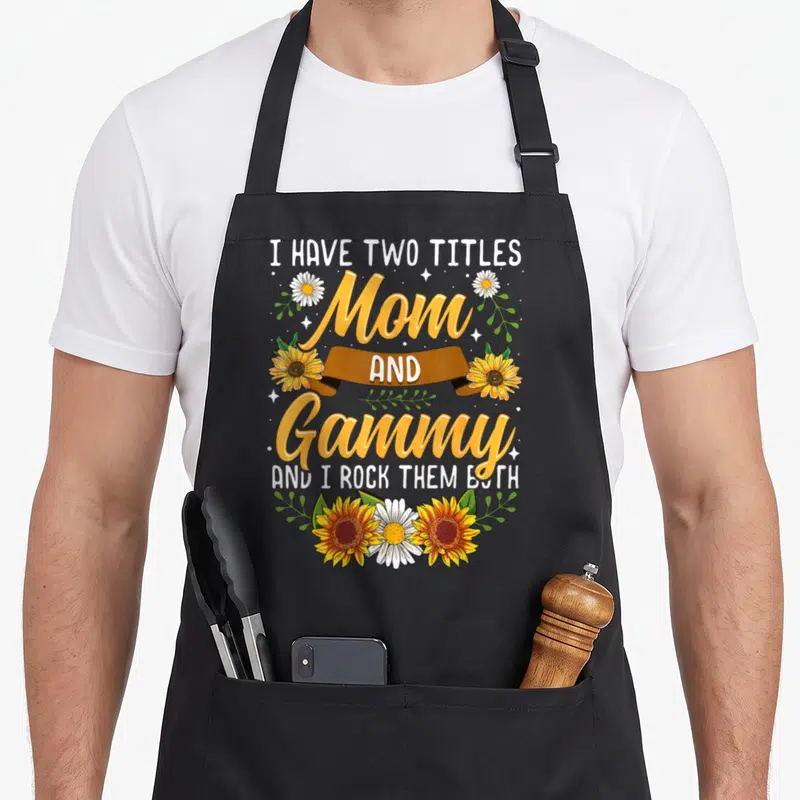 Title grandma co apron - Orange Mom and Gammy text with sunflowers daisies and brown ribbon — shown on a black cotton canvas apron against a white background