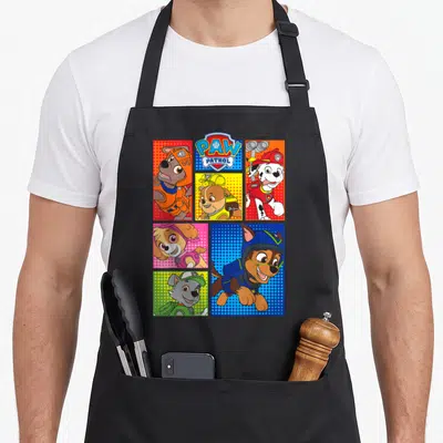 Pup novelty apron - Five panel grid of cartoon puppies with PAW PATROL logo in blue red yellow and orange — shown on a black cotton canvas apron against a white background